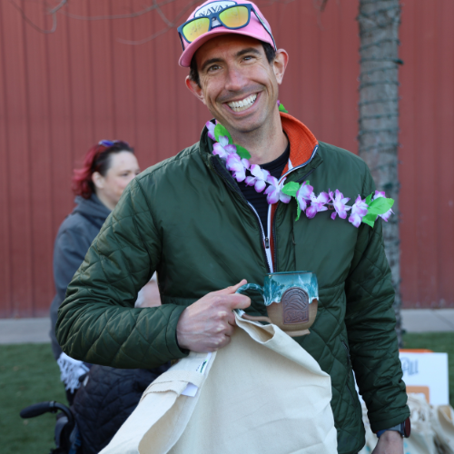 A person smiles toward the camera while holding a ceramic mug and canvas tote bag outdoors in front of a red building. They wear a pink hat with sunglasses on top, a green jacket, and a purple flower lei. Another person and tables with tote bags appear blurred in the background.