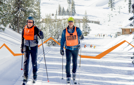 Two people cross-country ski side by side on a groomed trail, each using poles and moving forward with steady strides. One wears an orange vest that reads “VISUALLY IMPAIRED,” and the other wears an orange vest with O-A-S branding. Snow-covered trees, buildings, and skiers appear in the background, with an orange graphic line overlaying the scene.
