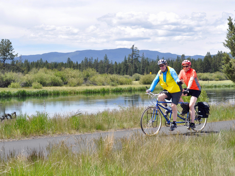 A tandem bike moves along a paved path beside a calm river; two people pedal together, one in front and one behind, both wearing helmets. Grassy banks, trees, and distant mountains sit under a partly cloudy sky in the background.