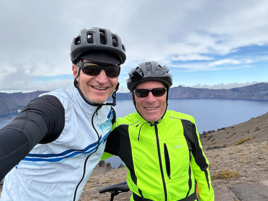Two people smile at the camera in a close-up selfie while wearing bike helmets and sunglasses, standing on a rocky overlook above a large lake. Steep crater walls and water fill the background under a cloudy sky, with part of a bike visible at the edge of the frame.