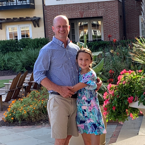 Kendall stands with hs daughter, in landscaped courtyard as she hugs his waist and they both smile toward the camera. Kendall wears a blue checkered button-up shirt, khaki shorts, and flip-flops. The girl wears a colorful floral dress and white sandals. 