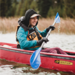 Kellie Standish paddles a red kayak across calm water, smiling as she holds a blue double-bladed paddle. She wears a teal waterproof jacket, a black hood over a light-colored cap, black gloves, and an orange P-F-D. Water droplets fall from the raised paddle blade, and reeds and evergreen trees line the far shore in the background.