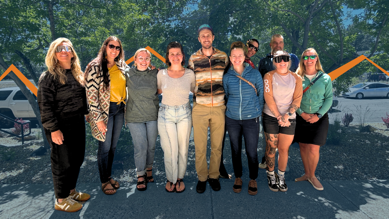 Melodie Buell, Charlotte Dadabay, Emma Flynn, Hayley McClure, Jesse Brown, Cara Frank, Taylor Cowan, and Carly Schmidt stand shoulder to shoulder in the front row, smiling toward the camera. Behind them, Pat Addabbo and Jared Apjoke stand partially visible between the group. Melodie wears a black long-sleeve outfit with tan sneakers and mirrored sunglasses. Charlotte wears a patterned cardigan over a yellow top, dark jeans, sandals, and sunglasses. Emma wears pink-framed glasses, a green striped top, cuffed jeans, and black sandals. Hayley wears a light gray T-shirt, light blue jeans, and flip-flops. Jesse wears a striped fleece jacket, tan pants, black shoes, and a baseball cap. Cara wears a blue puffy jacket, dark pants, and brown shoes. Taylor wears a tan and pink long-sleeve jersey, black shorts, tall patterned socks, black sneakers, large visor-style sunglasses, and has visible tattoos on one leg. Carly wears a green zip-up jacket, a black skirt, tan slip-on shoes, and green sunglasses. An orange line styled like a mountain ridgeline runs horizontally behind the group, weaving up and down across the image.