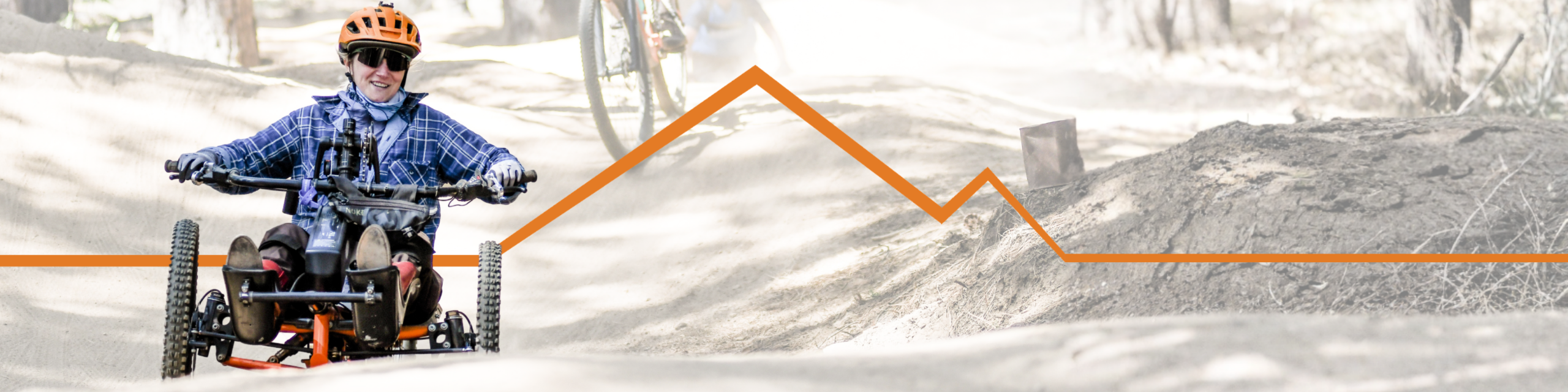 A person rides an adaptive mountain bike along a dusty trail through a forest. Another cyclist follows behind, and an orange mountain graphic overlays the image.
