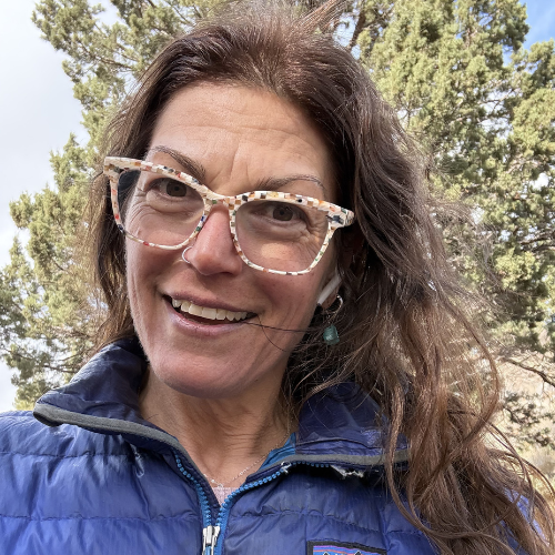 Leah, a white woman with shoulder-length brown hair, smiles at the camera while wearing checkerprint glasses