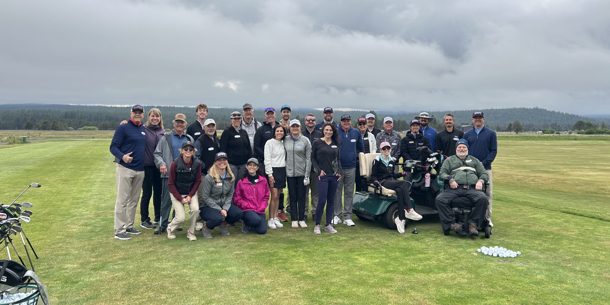 A group of 27 people stands on the putting green at Meadows Golf Course in Sunriver. Most are standing, one person is seated in an adaptive golf cart, and another in a power wheelchair