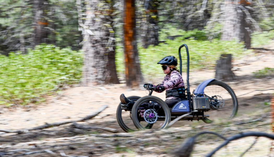 Ryan, wearing a pink and brown jersey, helmet and sunglasses rides an adaptive off-road handcycle along a dirt trail through a forest, with trees and green undergrowth blurred in the background.