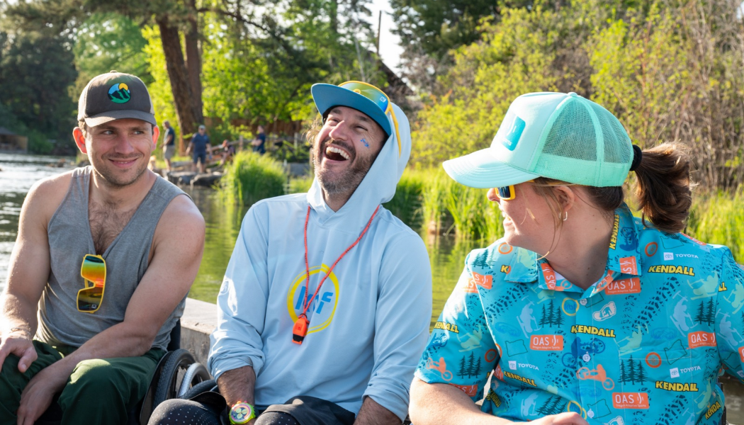 Three people seated in wheelchairs laugh and chat together on a dock beside a river. One wears a gray tank top, another a light blue hoodie, and the third a turquoise O-A-S shirt and matching hat. Trees and greenery line the riverbank behind them.