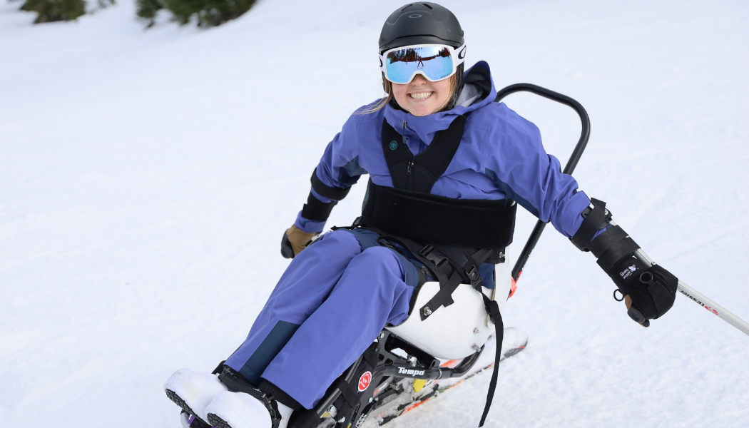 A smiling Ryan wearing a purple snowsuit, black helmet, and reflective blue goggles skis in a sitski on a snowy slope. She is secured in the sitski with black straps and holds outriggers in her hands.